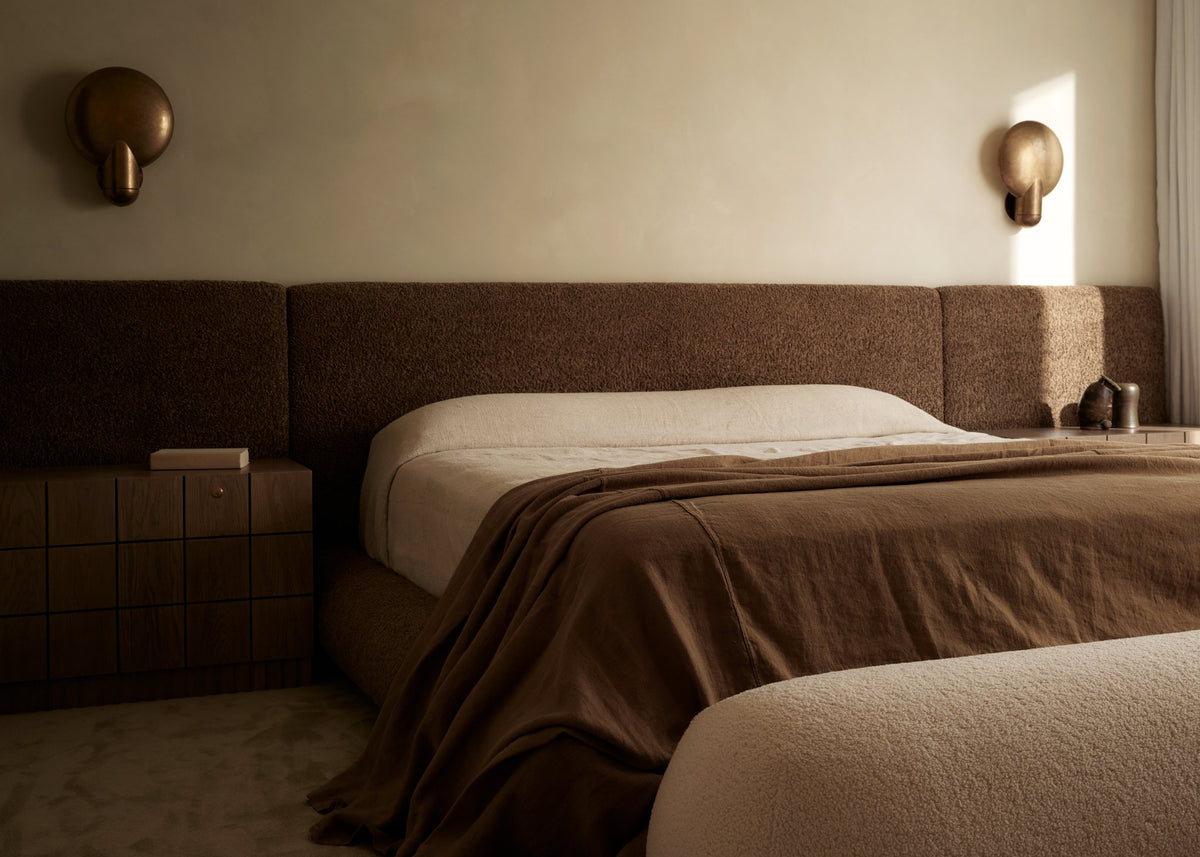 Bedroom with a wide upholstered headboard, layered brown linen bedding and sculptural wall sconces