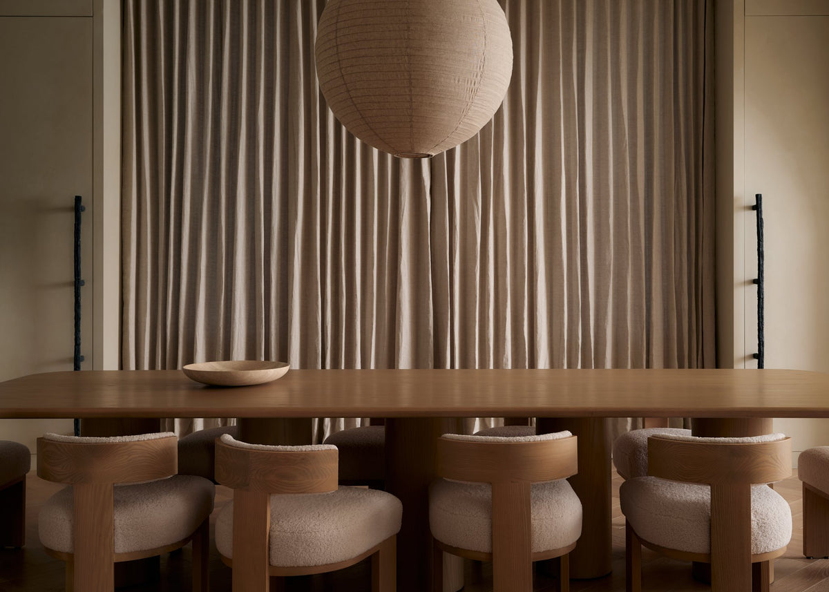 Dining room with a long oak table, curved upholstered chairs, a paper globe pendant and linen drapery