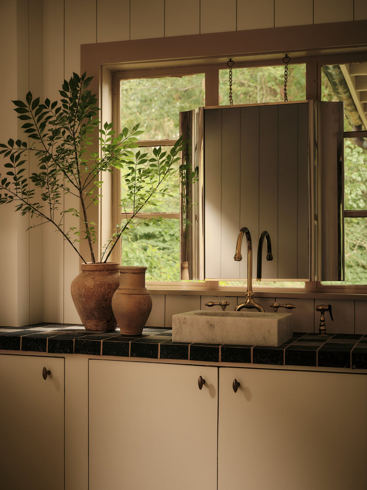 Kitchen with dark tiled counters, cream cabinetry, earthenware vessels and olive branches by the window