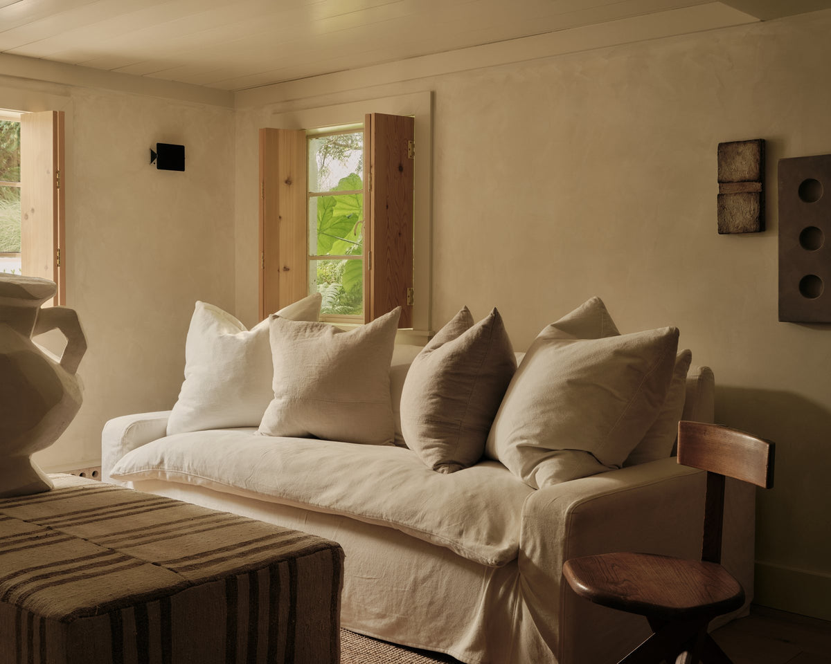 A sunlit living room styled with a slipcovered sofa, linen pillows, striped ottoman and sculptural vase