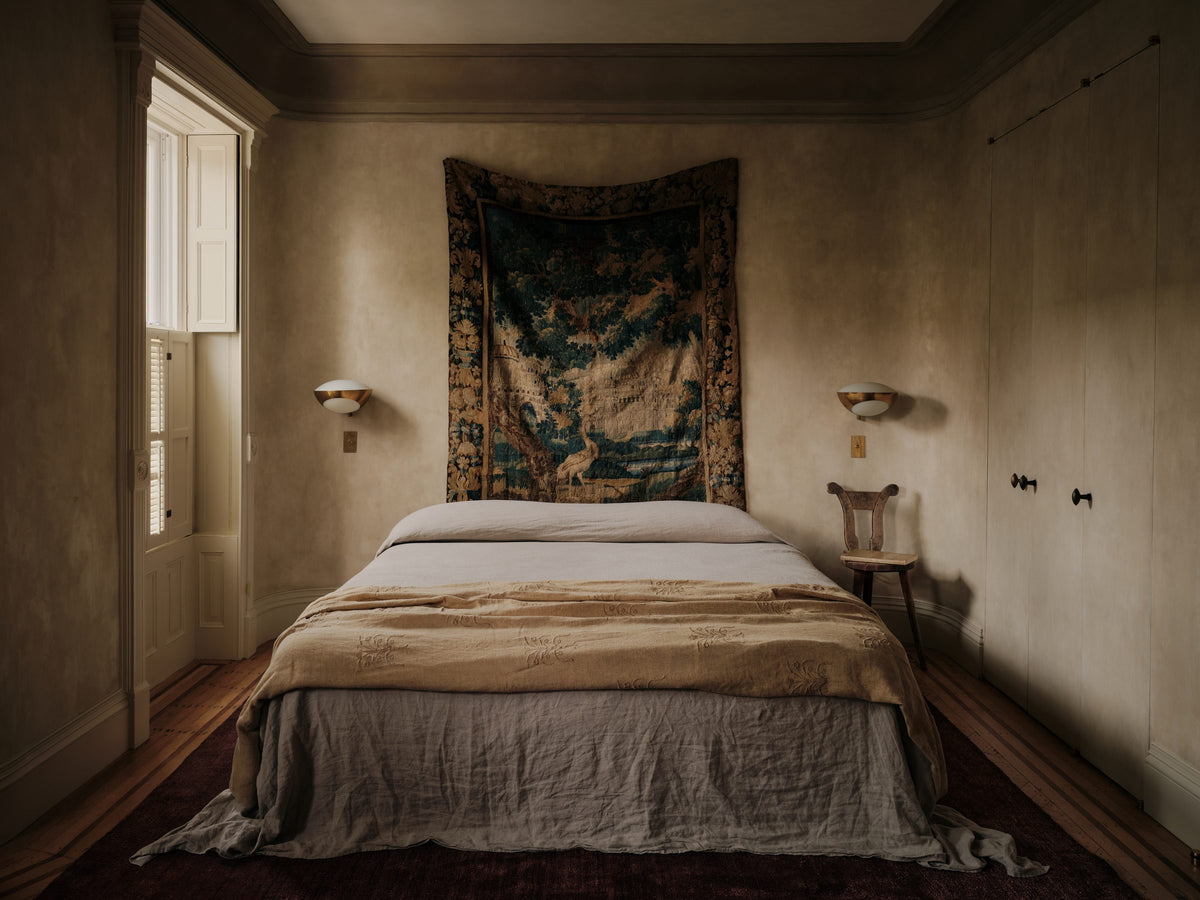 A calm bedroom with plaster walls, a linen-dressed bed and a woven tapestry above it
