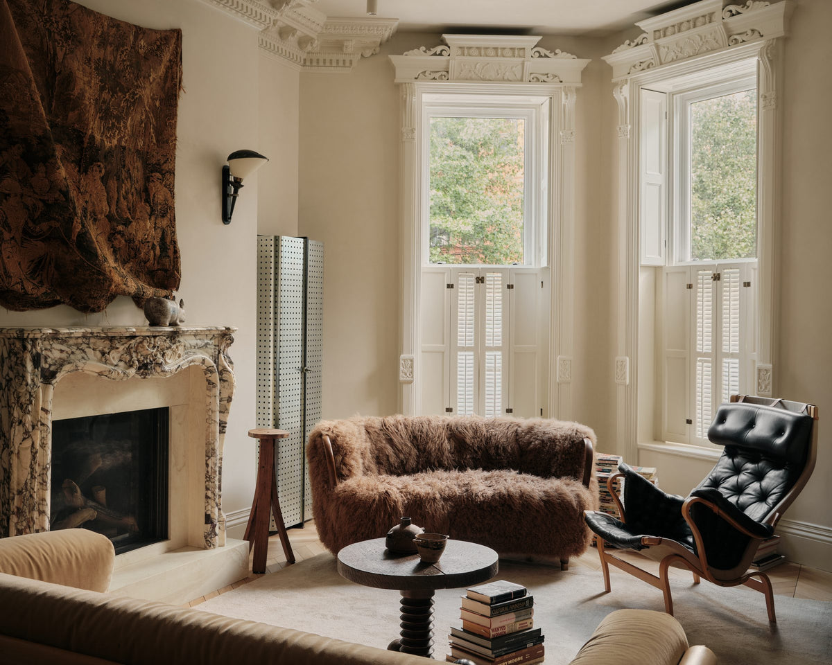 A sunlit living room with plaster walls, a shearling sofa and tall shuttered windows