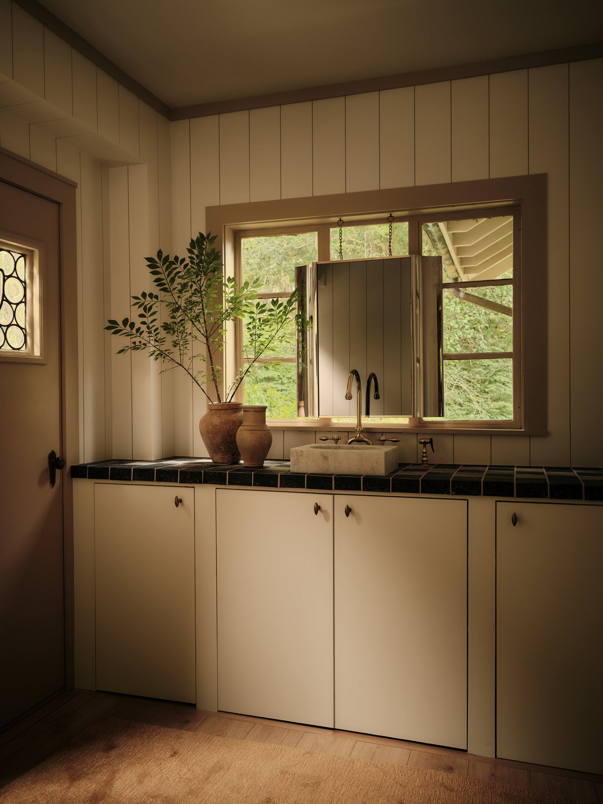 Cream kitchen cabinetry with round knobs, a dark tiled counter and stone sink beneath a garden window
