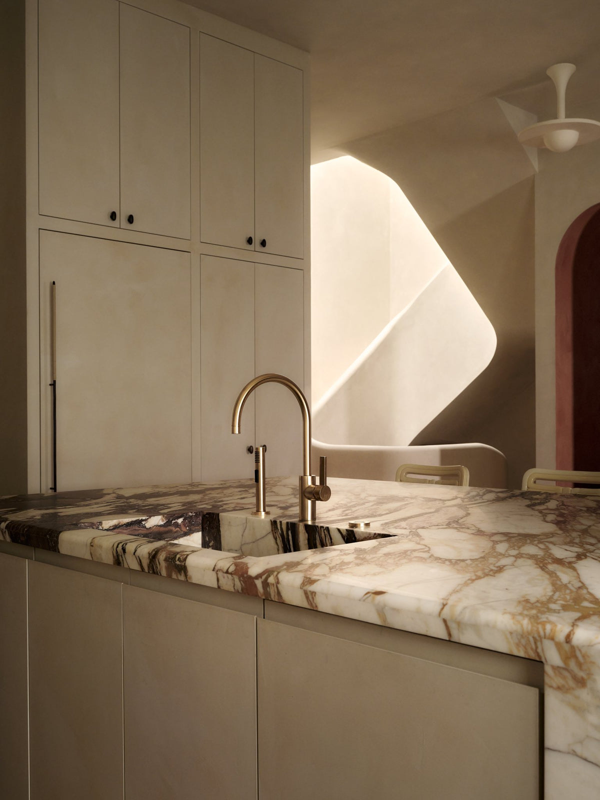 Kitchen with a veined marble island, brass faucet and cream cabinetry beside a curved plaster wall