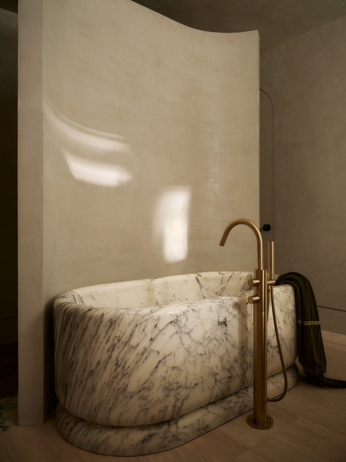 Freestanding marble bathtub with a brass floor-mounted tub filler against a curved plaster wall