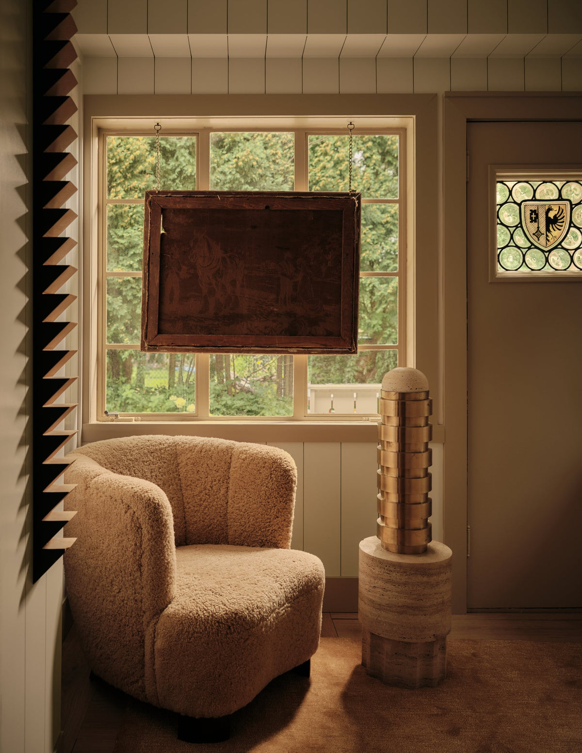 Shearling lounge chair beside a carved wood pedestal and garden window with vintage artwork